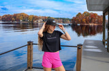 Woman standing on a dock by a lake, wearing a black t-shirt and pink shorts.